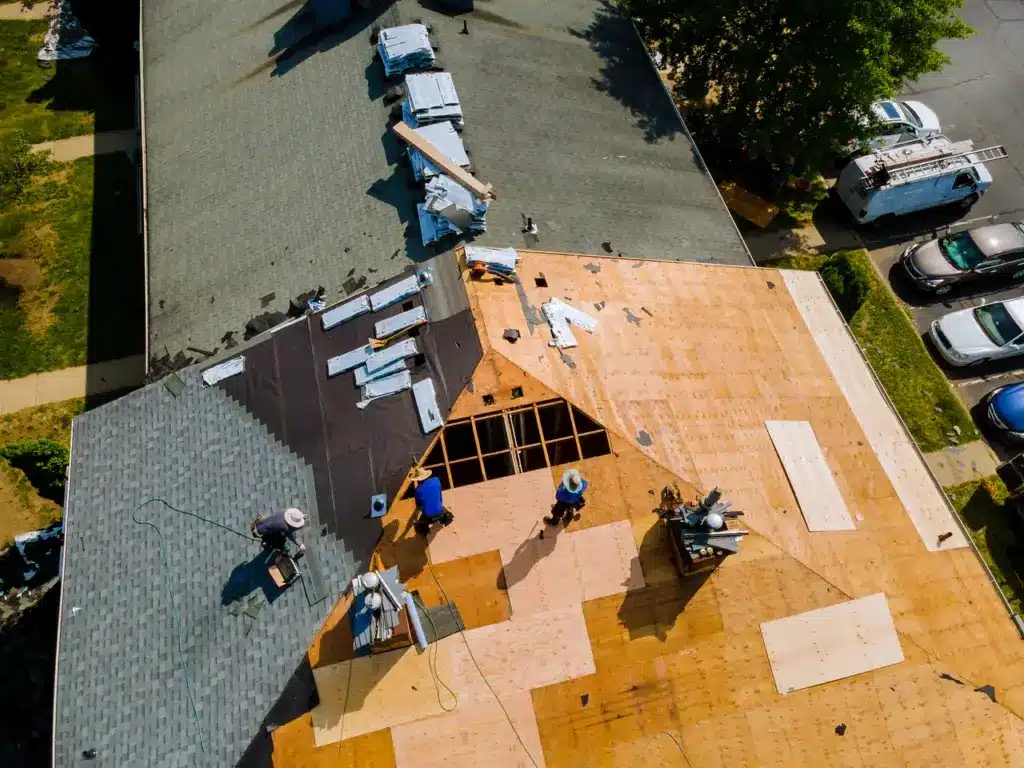 Aerial view of a residential roof under construction with exposed framing, plywood sheathing, and work crews.