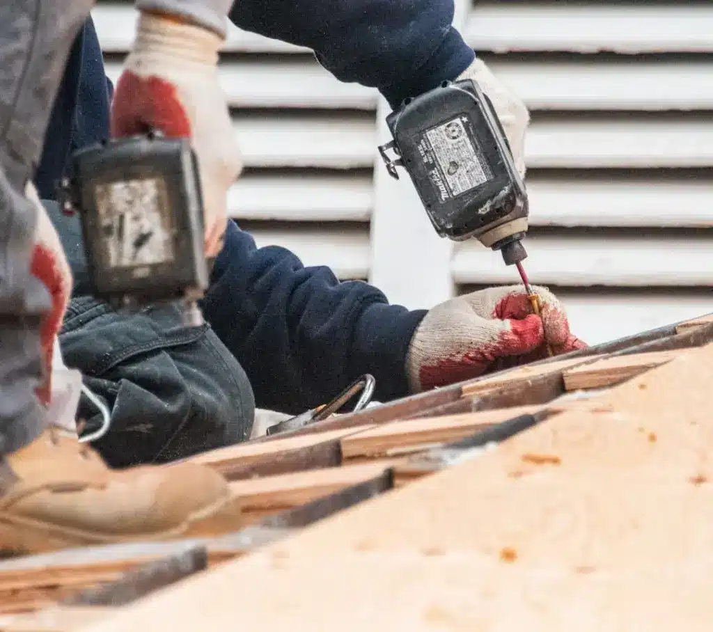 Worker in gloves using a pneumatic nail gun to fasten wood framing on a roof deck.