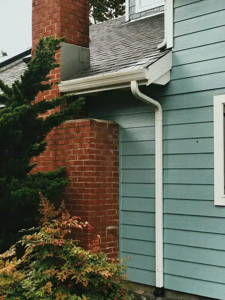 White downspout curves along green siding next to red brick chimney with ivy and shrubs.