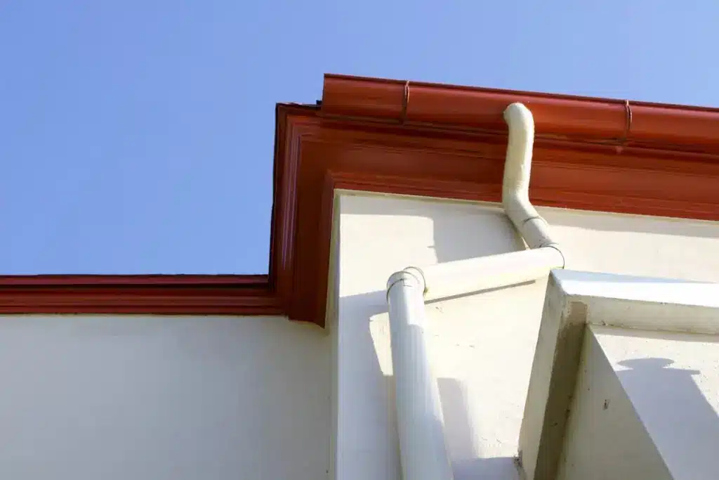 Red fascia board and white gutter system with downspout on residential roofline against blue sky.