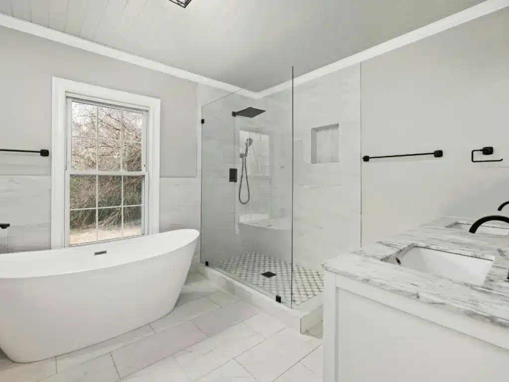 Modern bathroom with white marble countertops, freestanding tub, walk-in shower with glass enclosure, and large window.