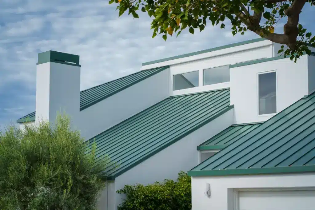 Contemporary white house with green metal roof panels and tall chimney, surrounded by landscaping.
