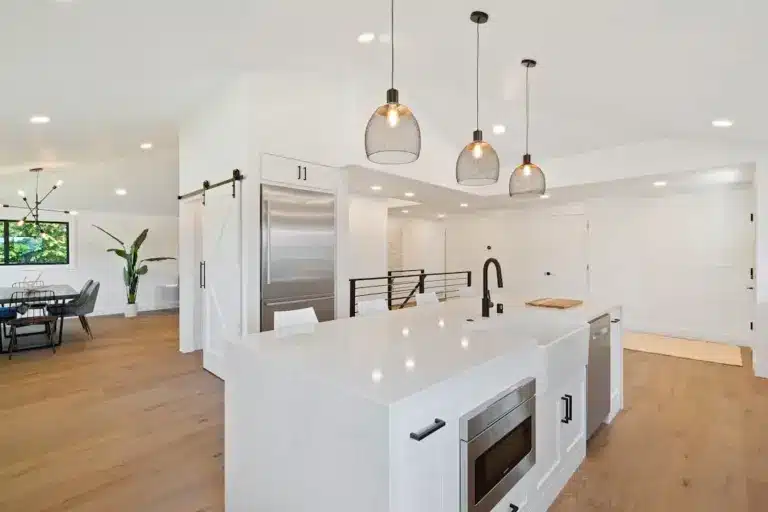 Modern kitchen with white island, stainless steel appliances, pendant lights, and hardwood floors.