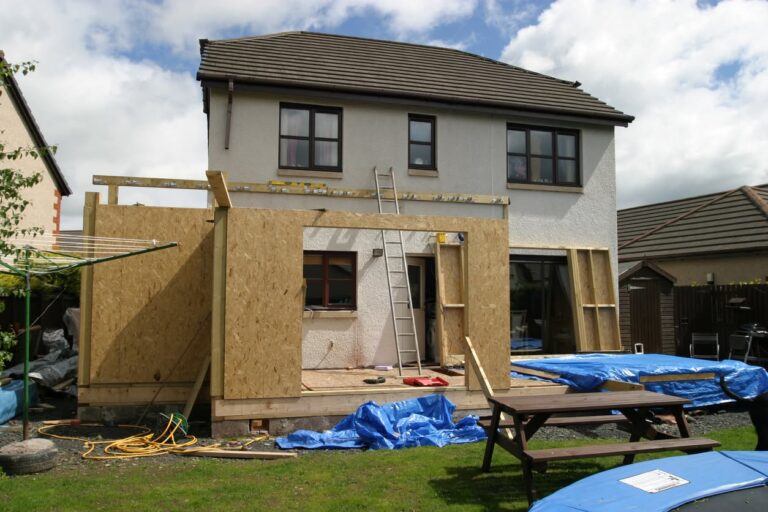 Rear of a two-story stucco house with a single-story addition framed in OSB panels, a ladder, blue tarps and a picnic table.