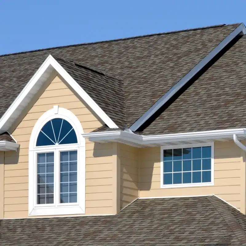 Close-up of residential roof with gray asphalt shingles, white trim, and gable window on cream-colored house.