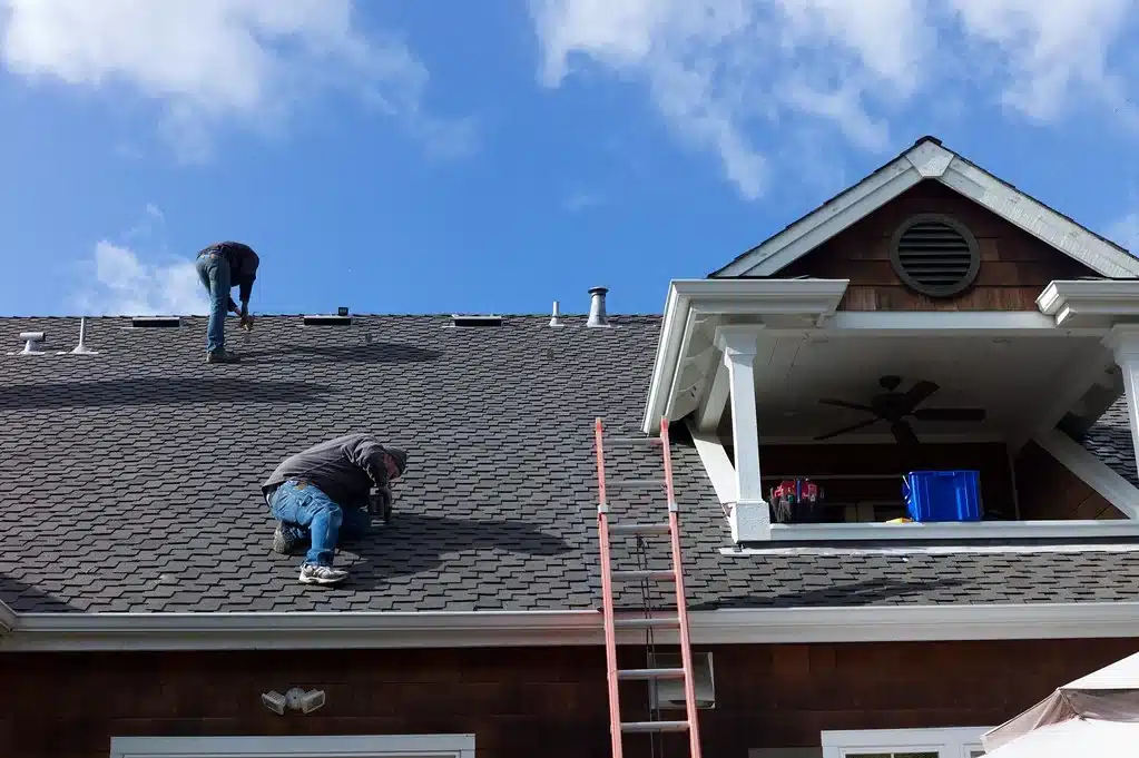 Two workers on a residential roof performing repair work on dark asphalt shingles.
