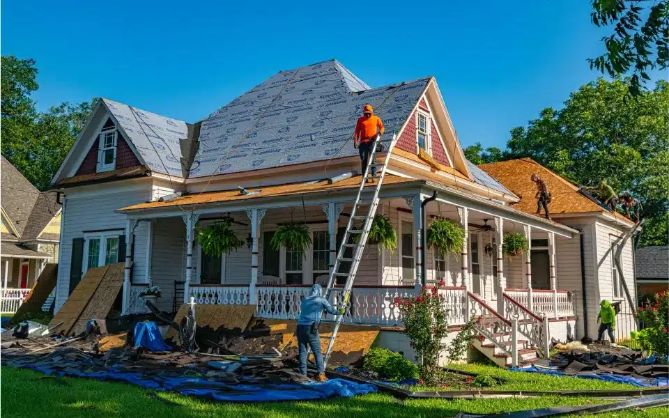 Worker on ladder installing blue underlayment on pitched roof of Victorian-style house during renovation.