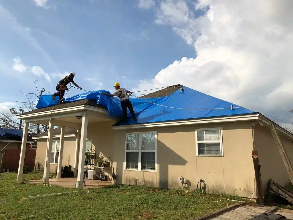 Two workers on a residential roof securing blue tarps to protect the house during repairs.