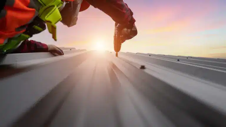 Worker in safety gear installing metal roofing panels at sunrise with corrugated sheets visible.