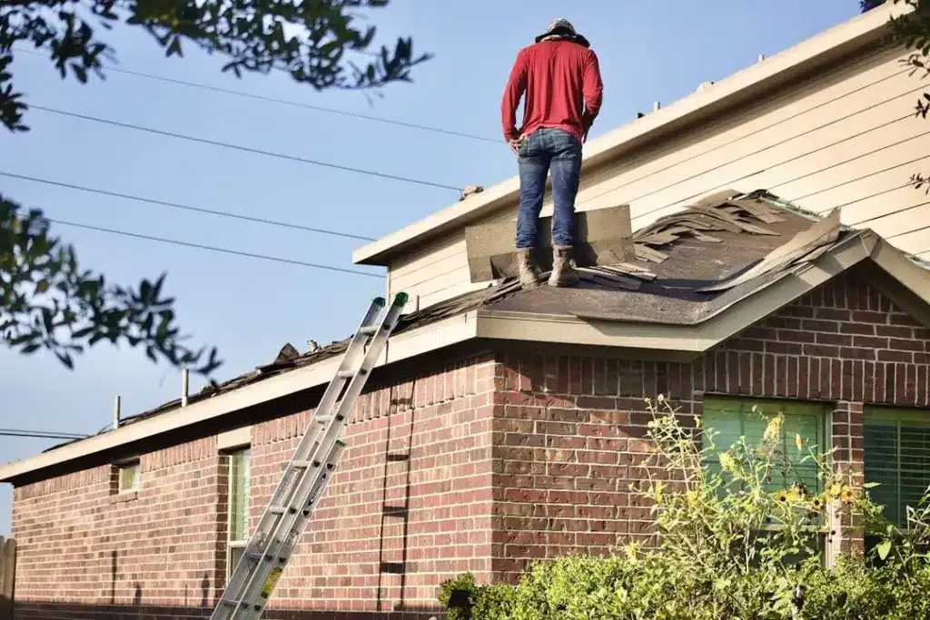 Worker in red jacket standing on roof of brick home next to ladder, removing damaged shingles.