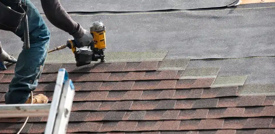 Roofer using pneumatic nailer to install asphalt shingles on residential roof.