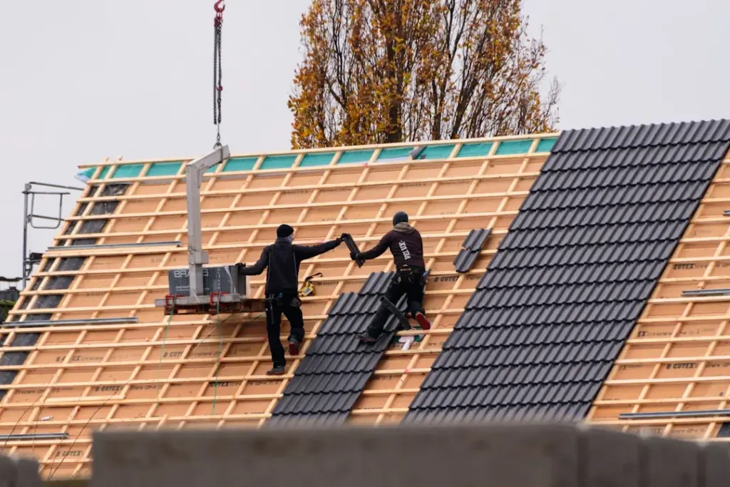 Two roofers in safety gear installing dark shingles on a sloped roof with exposed wooden framing and underlayment.