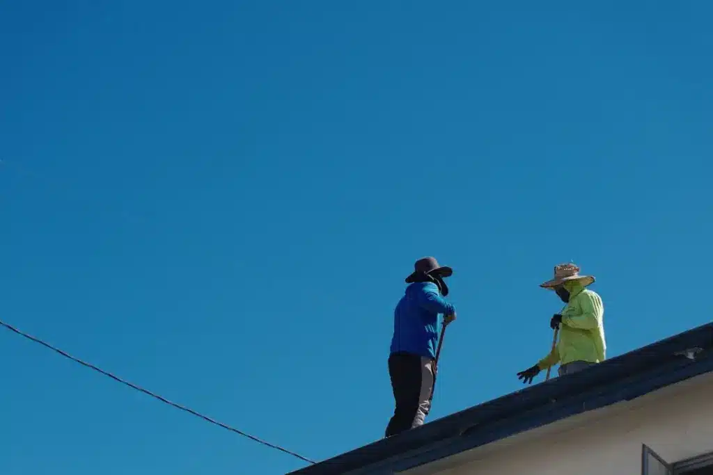 Two roofers stand on a residential roof edge against a clear blue sky.
