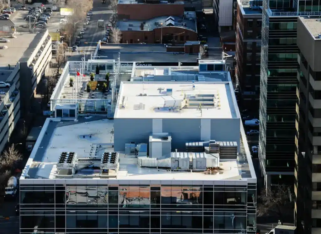 Aerial view of a commercial rooftop with HVAC equipment, mechanical systems, and white safety railings surrounded by urban buildings.