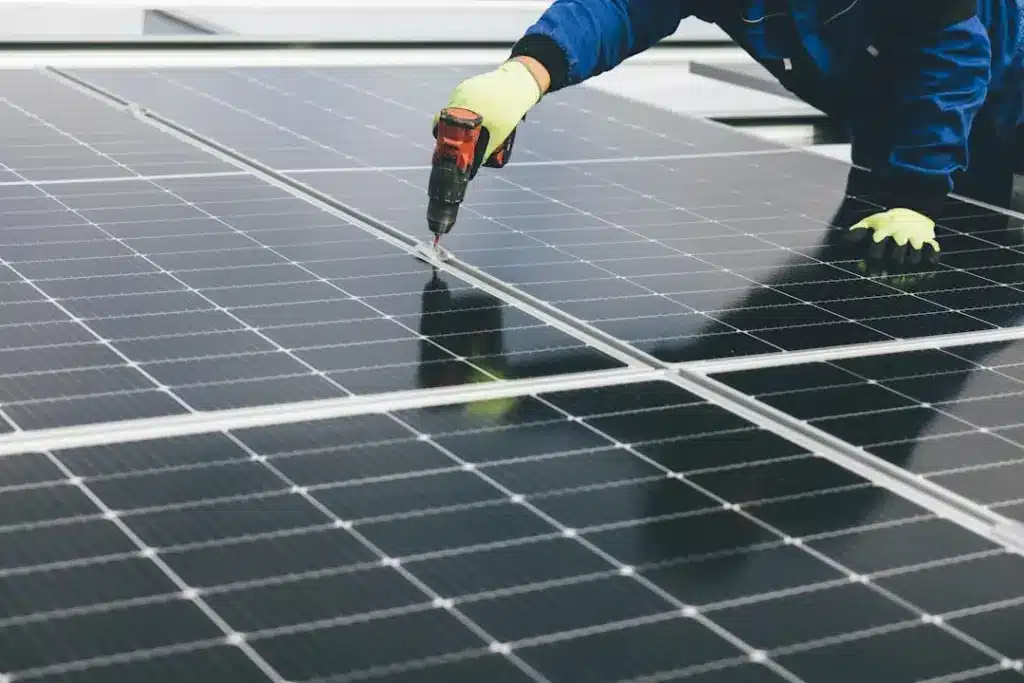 Worker in blue jacket and yellow gloves using power drill to secure solar panel mounting rail.