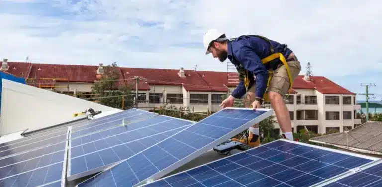 Roofer in safety gear installing solar panels on residential rooftop with apartment buildings in background.