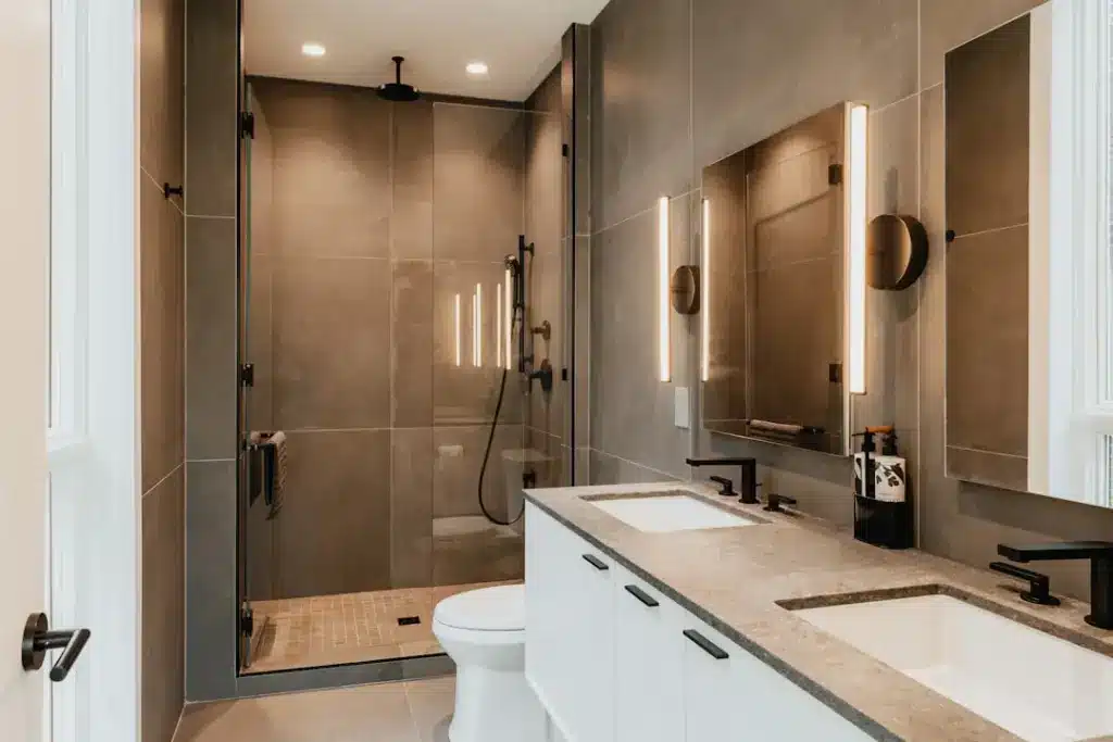 Modern bathroom with dual vanities, large mirror with lit sconces, walk-in shower with glass enclosure and dark fixtures.